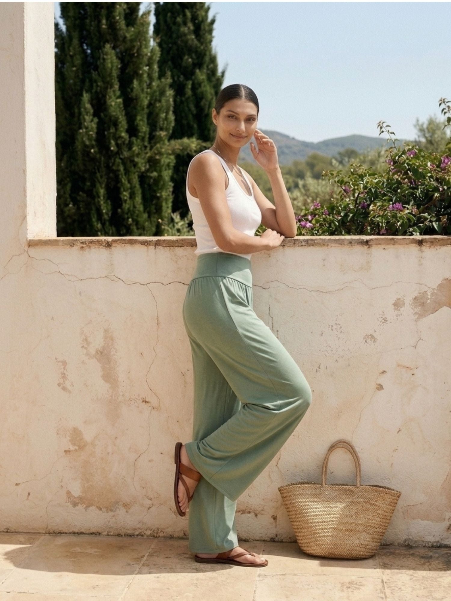 Woman in sage green wide-leg pants and white tank top, leaning on wall with woven tote bag, ideal for travel and everyday wear.