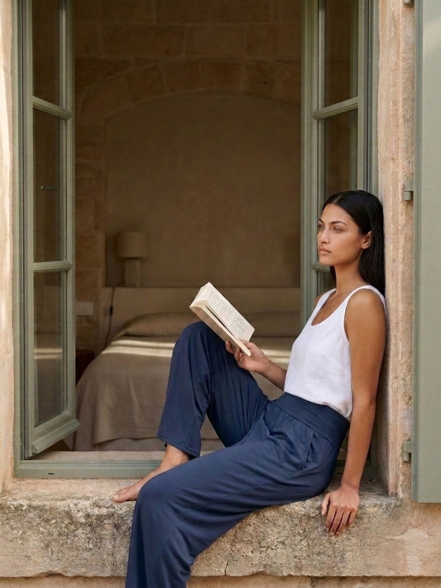 Woman relaxing by window with book, wearing soft sustainable travel lounge pants and tank top